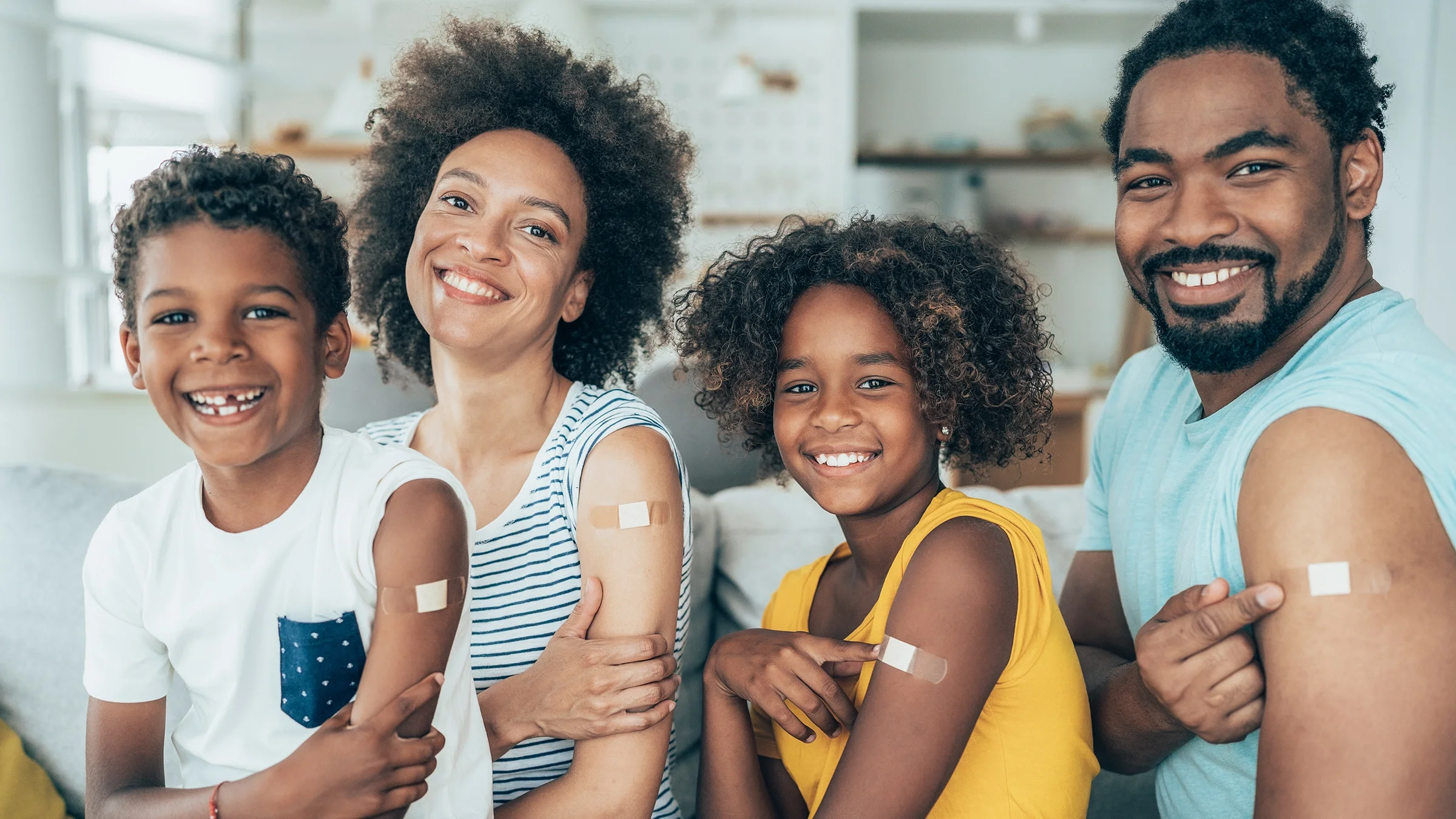 Family of four showing off their band-aids from the COVID-19 vaccine. They are sitting on their couch at home.