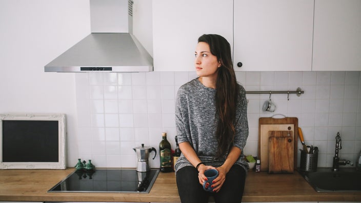 A woman sits alone in the kitchen, holding a cup of tea.