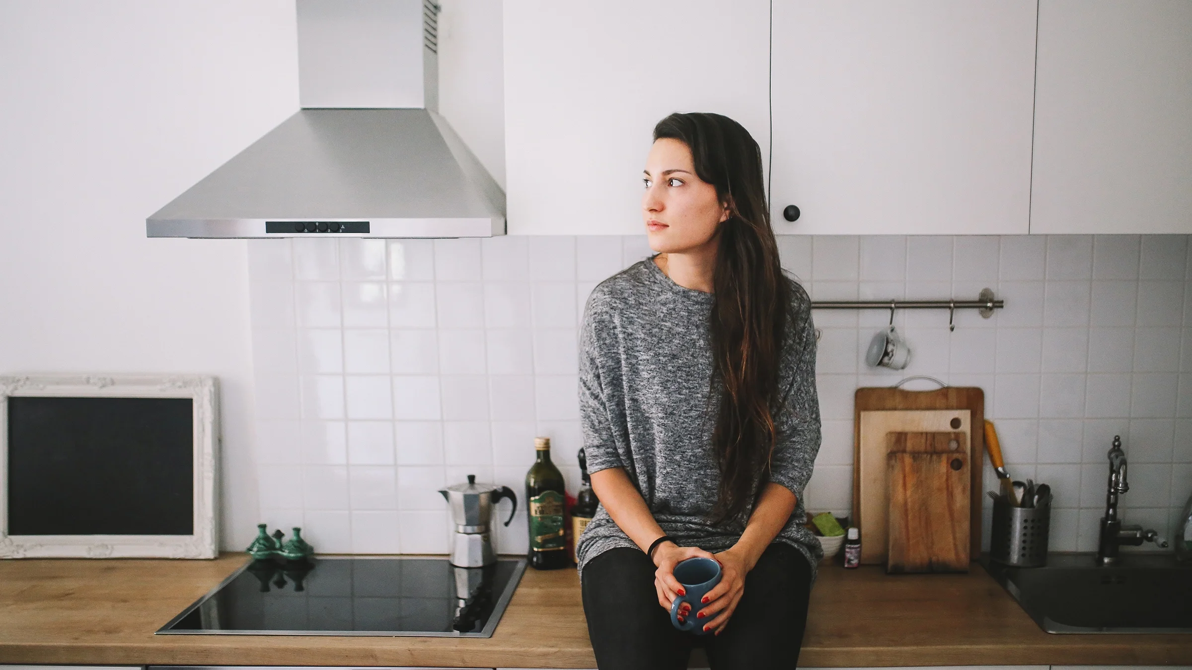 A woman sits alone in the kitchen, holding a cup of tea.