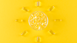 Vitamin D capsules in a clear dish in the middle of the frame surrounded by individual capsules creating a sunray pattern around the dish on a bright yellow background.
nadisja/iStock via Getty Images