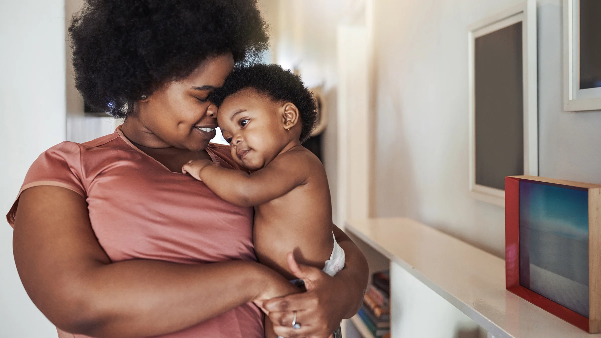 Portrait of a mother holding her baby close. She is wearing a pink t-shirt and nuzzling close and kissing her baby's forehead.