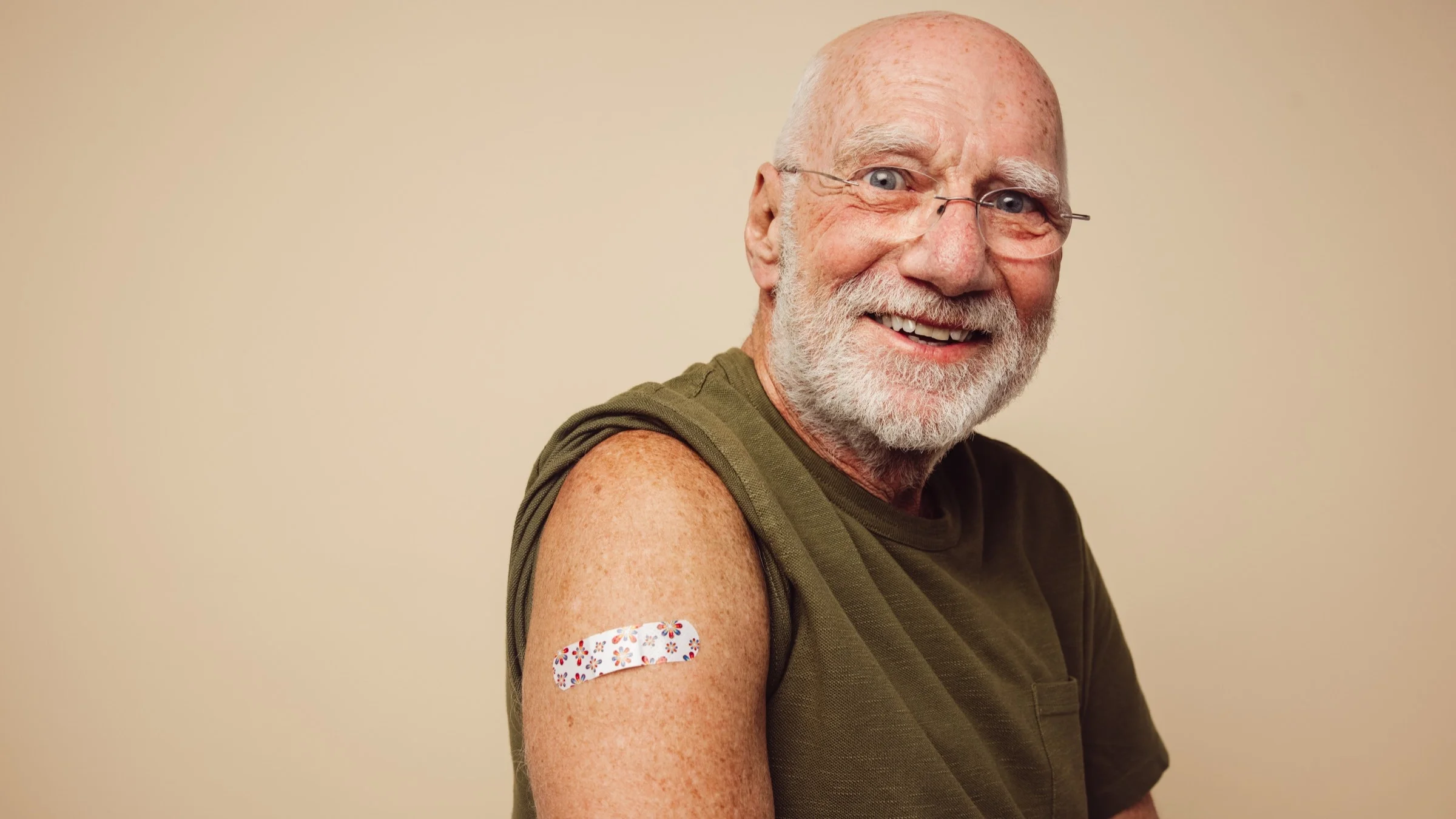 Portrait of an older man with his sleeve rolled up to show his flower band-aid off after getting a vaccine on a tan background.