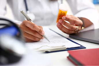 Healthcare professional writing on a clipboard while holding a prescription bottle at a desk